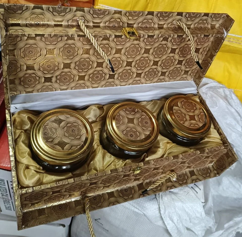 A gold patterned box with rope handles, holding three glass jars with gold-tone patterned lids on gold satin