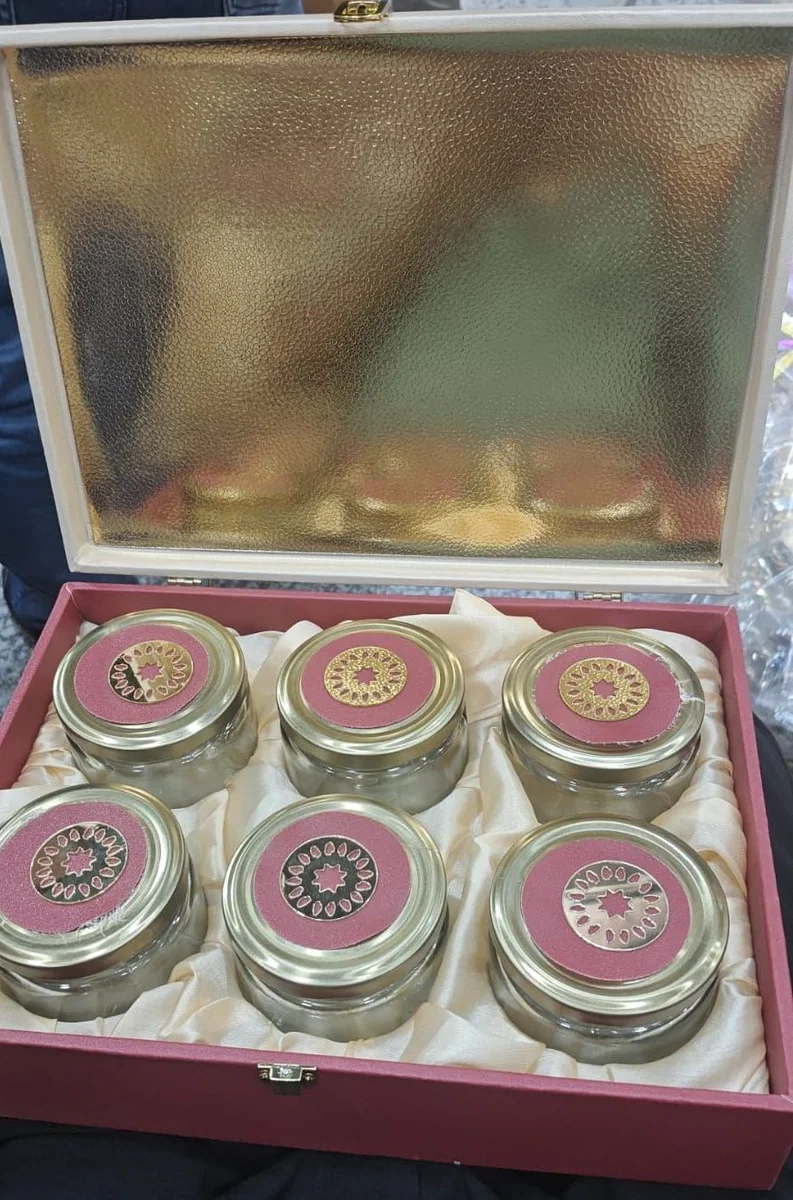 A red box with a gold-lined lid, holding six glass jars with rose and brass-tone lids on cream satin