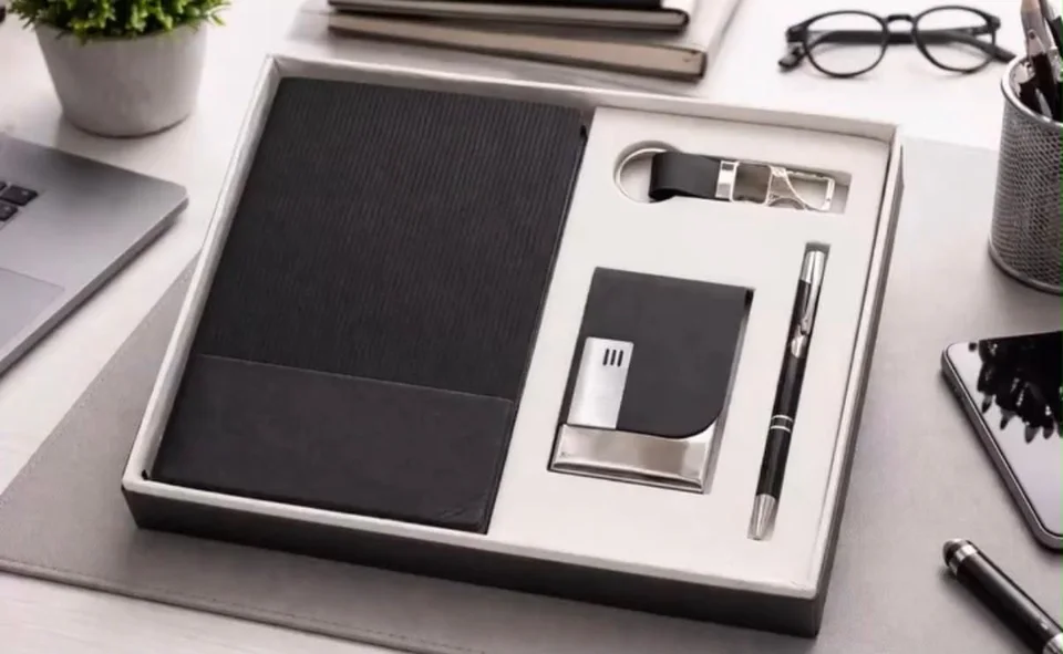 A black notebook with a ribbed cover, a black card holder with a chrome edge, a black pen and a black strap keychain, in a…